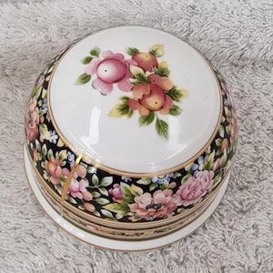 Puede incluir: Una caja de baratijas redonda de porcelana con tapa. La tapa presenta un diseño floral con flores rosas y melocotón y hojas verdes. Los lados de la caja están adornados con un fondo negro y un estampado floral.
