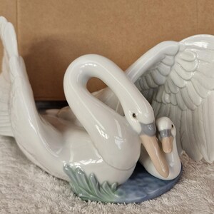 May include: A white ceramic swan figurine with a smaller swan nestled beside it. The larger swan has outstretched wings and a curved neck, with a light blue base and green accents. The figurine is a decorative item.