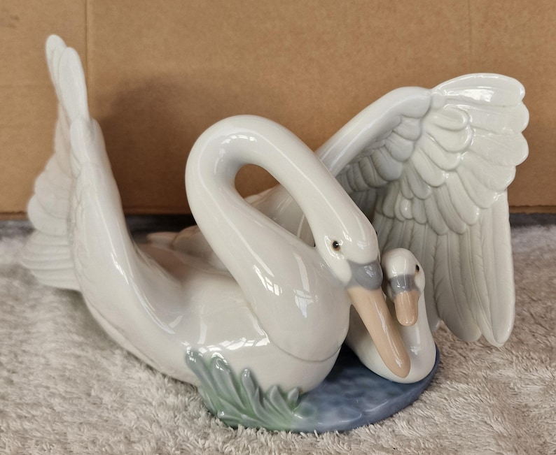May include: A white ceramic swan figurine with a smaller swan nestled beside it. The larger swan has outstretched wings and a curved neck, with a light blue base and green accents. The figurine is a decorative item.