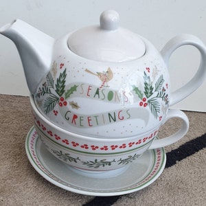 May include: White ceramic teapot and cup set with a festive holiday design. The teapot and cup feature a green and red holly design with a red bird and the text "Seasons Greetings".