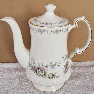 May include: White porcelain teapot with a floral design featuring purple and white flowers. The teapot has a gold trim around the lid and base.