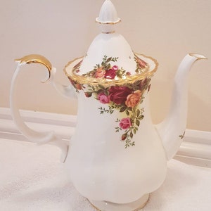 May include: White porcelain coffee pot with a floral rose pattern in red, pink, and yellow. The pot has a gold handle and trim, and a matching lid with a finial. The spout is curved. The pot is decorated with roses.