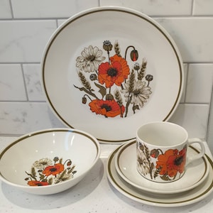 Könnte beinhalten: Ein Set aus weißem Keramikgeschirr mit einem Blumenmuster aus roten Mohnblumen, weißen Gänseblümchen und grünen Weizenähren. Das Set enthält einen Speiseteller, eine Schüssel und eine Teetasse mit Untertasse.
