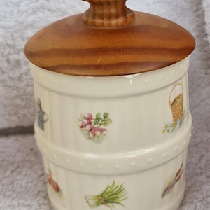 May include: A white ceramic jar with a wooden lid. The jar has a barrel design and is decorated with small floral and vegetable illustrations. The lid has a knob on top.
