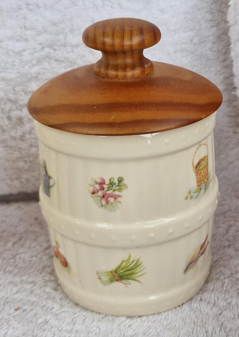 May include: A white ceramic jar with a wooden lid. The jar has a barrel design and is decorated with small floral and vegetable illustrations. The lid has a knob on top.