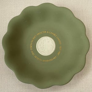 Puede incluir: Un plato de cerámica verde con un diseño en relieve blanco y letras doradas. El texto dice "Procter & Gamble Limited - 1837 - 1987 - And Hundred And Fiftieth Anniversary".