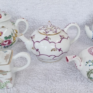 May include: Five miniature porcelain teapots with floral and geometric designs. The teapots are white with pink, purple, and gold accents.