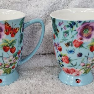May include: Two light blue ceramic mugs with floral and butterfly patterns. The mugs have light blue handles and bases, and white interiors. The floral design includes red strawberries, purple flowers, and butterflies. A ladybird is on the base.