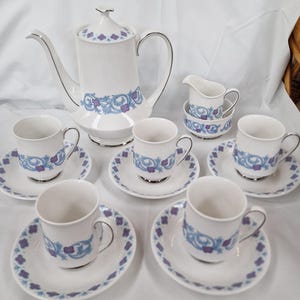 Könnte beinhalten: Ein weißes Porzellan-Kaffeeservice mit blauem und lila Blumendesign. Das Set umfasst eine Kaffeekanne, einen Sahnekännchen, eine Zuckerdose und sechs Tassen mit Untertassen. Silberne Verzierungen säumen die Ränder der Teile.