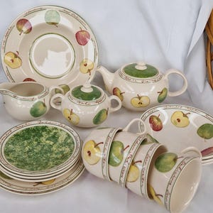 May include: A ceramic tea set featuring a cream-coloured base with hand-painted apple illustrations in red, green, and yellow. The set includes a teapot, sugar bowl, creamer, stacked cups, and plates, all decorated with cursive text naming apple varieties.