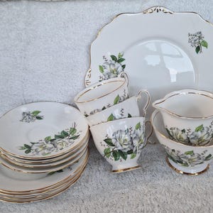 Könnte beinhalten: Ein Vintage-Teeservice mit weißen Porzellanstücken mit einem floralen Design in Grau und Grün. Das Set umfasst Tassen, Untertassen, einen Sahnekännchen, eine Zuckerdose und einen großen Servierteller, alle mit Goldrand.