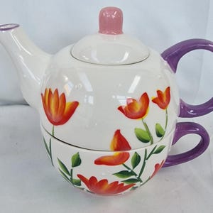May include: A white ceramic teapot and teacup set with a floral design. The teapot has a purple handle and spout, a pink lid, and is decorated with orange and red flowers and green leaves. The teacup has a purple handle and matches the teapot's design.