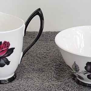 Puede incluir: Un juego de jarra de crema y azucarero blanco con ribete negro y un diseño de rosa negra y roja.