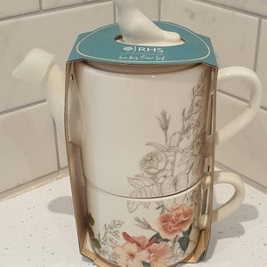 May include: Cream-colored tea for one set with floral design. The set includes a teapot with a bird-shaped lid and a matching teacup. The floral design features pink and beige roses and foliage. The packaging reads "tea for One Set".