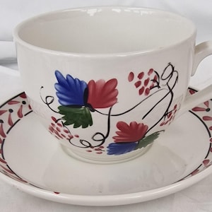 Puede incluir: Un juego de taza y plato de cerámica color crema con un diseño pintado a mano de hojas rojas y azules, follaje verde y pequeños racimos rojos. El borde del plato está decorado con un patrón repetitivo de pequeñas formas de hojas rojas.