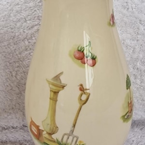 May include: A cream-colored ceramic vase with a bulbous body and a round base. The vase is decorated with a whimsical garden scene featuring a bird, a small tree, a fence, and a garden tool.