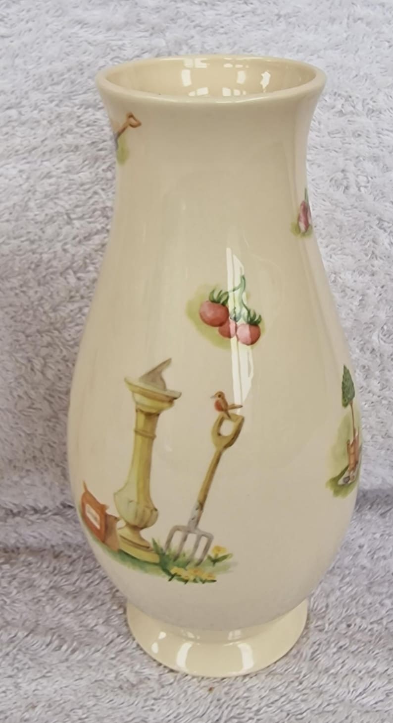 May include: A cream-colored ceramic vase with a bulbous body and a round base. The vase is decorated with a whimsical garden scene featuring a bird, a small tree, a fence, and a garden tool.