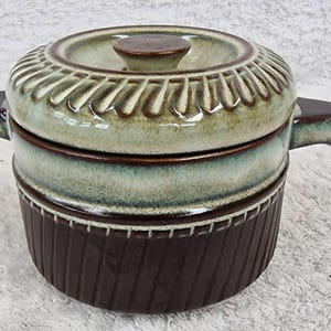 Puede incluir: Una cazuela de cerámica con tapa y esmalte marrón y verde. La tapa tiene un diseño decorativo y un pequeño pomo. El plato tiene dos asas y una base acanalada. Un artículo de cocina vintage.