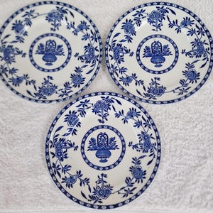 Puede incluir: Tres platos de cerámica blanca con diseños florales azules. Cada plato tiene un motivo central de cesta con un borde floral azul y blanco.