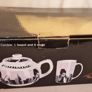 Könnte beinhalten: Eine schwarze Box mit einer weißen Teekanne und Tasse mit einem Stadtbild-Design. Die Teekanne trägt den Schriftzug "Manhattan". Die Box enthält außerdem den Text "Manhattan by Paul Cardew, 1 teapot and 4 mugs" und "Paul Cardew exclusive to ethos al".