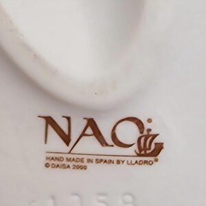 May include: Close-up of a white ceramic object with the brown text "NAC" and "HAND MADE IN SPAIN BY LLADRO". The text "DAISA 2000" is also visible. The object has a curved shape and a smooth surface.