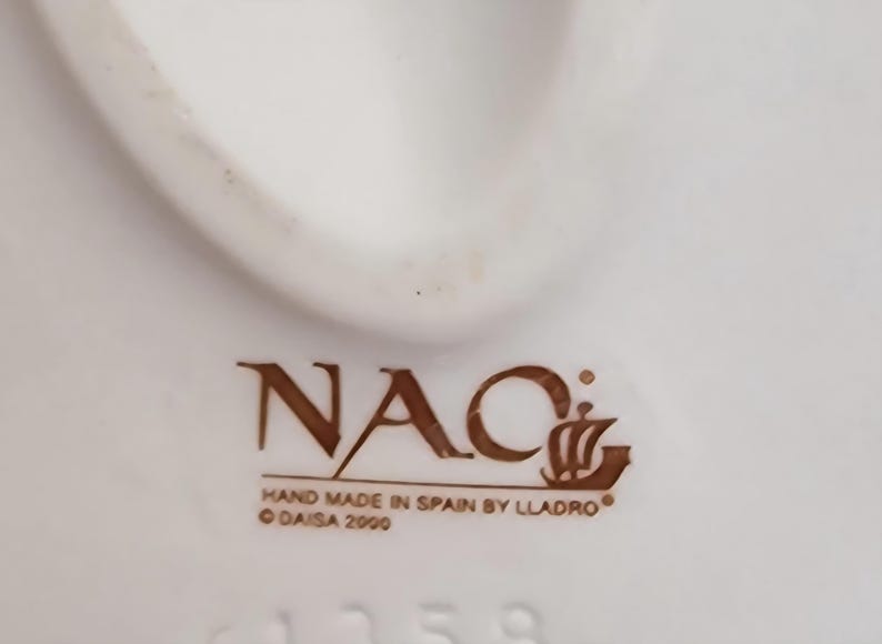 May include: Close-up of a white ceramic object with the brown text "NAC" and "HAND MADE IN SPAIN BY LLADRO". The text "DAISA 2000" is also visible. The object has a curved shape and a smooth surface.