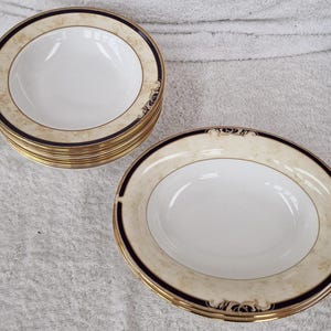 May include: A collection of white ceramic bowls with a cream border, navy blue trim, and gold detailing. The bowls have a classic design, suitable for serving soups or salads. The set includes several bowls.