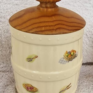 May include: A white ceramic canister with a wooden lid. The canister is decorated with a garden theme, including a rake, a hat, and a basket of fruit.
