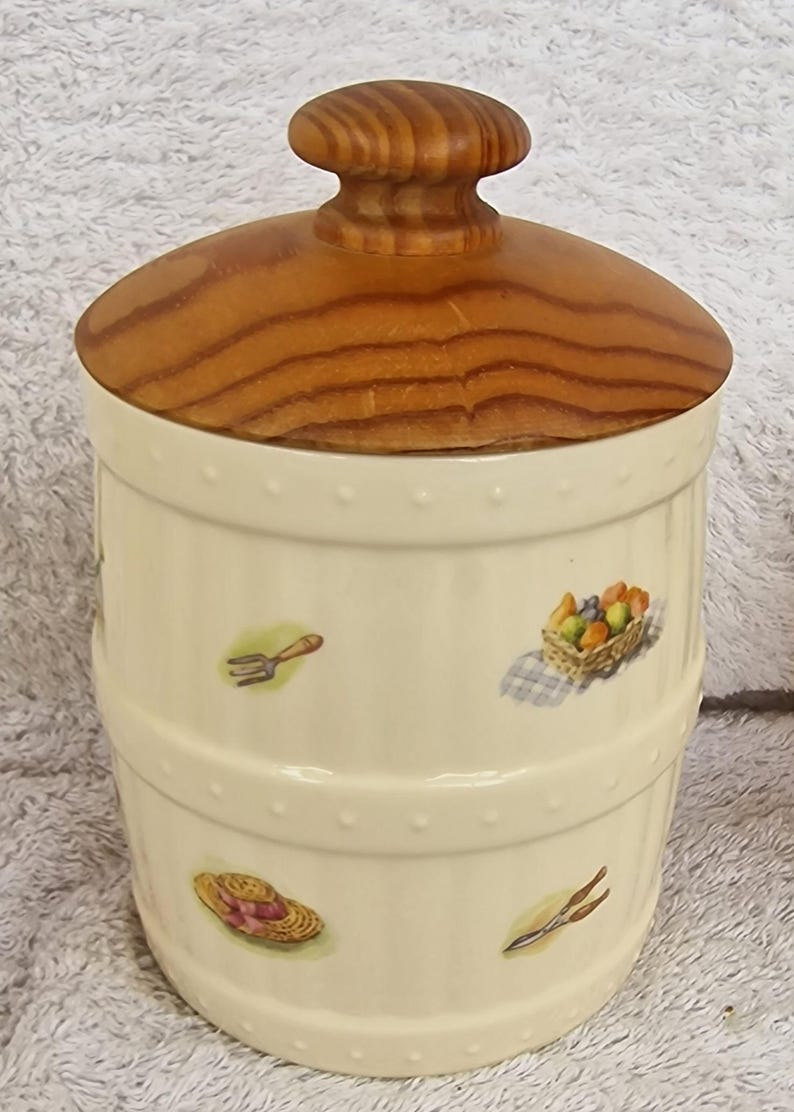 May include: A white ceramic canister with a wooden lid. The canister is decorated with a garden theme, including a rake, a hat, and a basket of fruit.