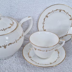 Könnte beinhalten: Ein weißes Teeservice mit goldenen Verzierungen. Das Set beinhaltet eine Teekanne, eine Milchkännchen, eine Zuckerdose, eine Tasse mit Untertasse und einen Teller.