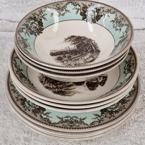 May include: A stack of vintage ceramic bowls and plates. The bowls have a light blue floral border and a black and white landscape scene in the center. The plates have a matching border. The set is stacked on a white surface.