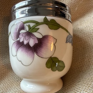 May include: A white porcelain egg coddler with a glossy finish, featuring a hand-painted purple and white flower design with green leaves and vines. The container has a polished silver-tone metal lid with a circular handle on top.