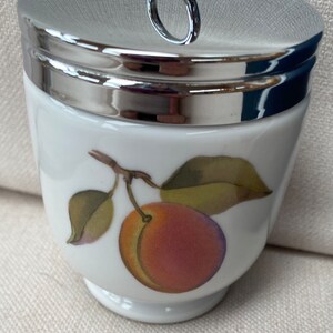May include: White ceramic egg cup with a silver lid and a painted image of a peach with green leaves.