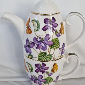 May include: A white porcelain tea set for one, featuring a stacked teapot and teacup design. The surface is decorated with purple violet flowers, green leaves, and orange and yellow butterflies. The set includes gold trim on the lid and rims.