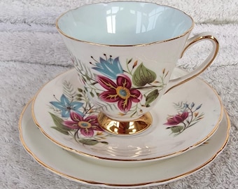 May include: A vintage teacup, saucer, and side plate set. The white porcelain features a floral design in burgundy, blue, and green, with gold trim. The teacup has a light blue interior and a gold-colored base.
