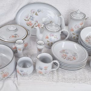 May include: A set of white ceramic dinnerware with a floral pattern. The set includes a large platter, a serving bowl, a teapot, a creamer, a sugar bowl, and several plates and bowls.