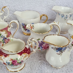 Peut inclure: Une collection de huit crémiers en porcelaine blanche avec des bordures dorées et des motifs floraux. Les crémiers ont des motifs floraux différents, notamment des roses roses, des fleurs violettes et des fleurs bleues.