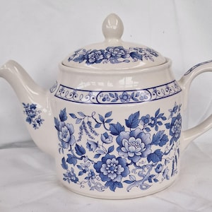 Puede incluir: Tetera de cerámica color crema con tapa, adornada con motivos florales azules. La tetera presenta un pico curvo, un asa redondeada y una banda decorativa alrededor del borde. La tapa tiene un pequeño pomo para facilitar la extracción.