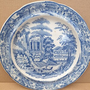 May include: A blue and white porcelain plate with a scenic design. The plate features a blue and white landscape with a castle, trees, a river, and a bridge. The border of the plate is decorated with a repeating pattern of houses and trees.