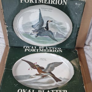 Pode incluir: Duas travessas ovais Portmeirion nas suas caixas originais. As travessas apresentam ilustrações de aves, uma com uma gansa-de-faces-brancas e outra com um tordo-ruivo. As caixas são verdes com texto branco, incluindo "PORTMEIRION" e "OVAL PLATTER".