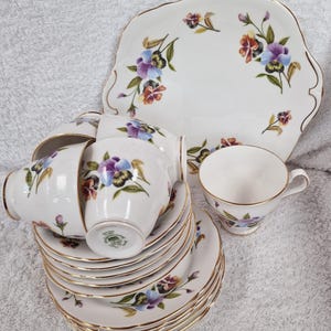 Può includere: Un set di tazze da tè e piattini vintage con un motivo floreale. Le tazze e i piattini sono bianchi con un bordo dorato e presentano un delicato motivo floreale con fiori viola, blu e gialli. Il set include un piatto da torta abbinato.