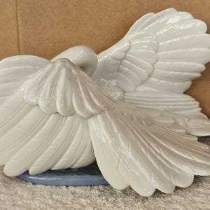 May include: A white ceramic swan figurine with outstretched wings. The swan is resting on a light blue base. The wings are detailed with textured feathers, and the swan's head is tucked down.