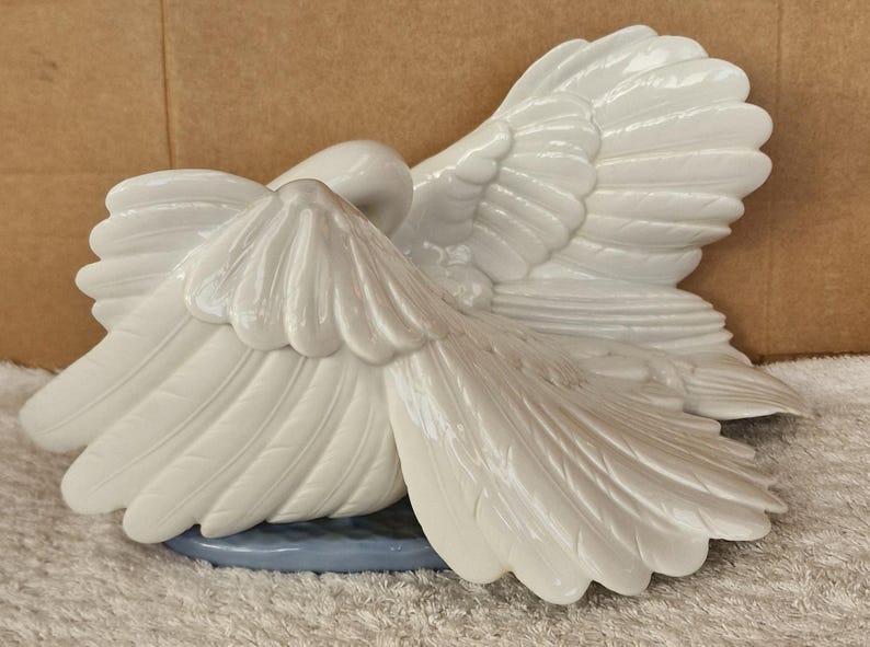 May include: A white ceramic swan figurine with outstretched wings. The swan is resting on a light blue base. The wings are detailed with textured feathers, and the swan's head is tucked down.