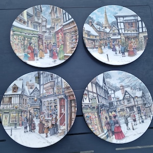 May include: Four round ceramic collector plates featuring detailed winter village scenes with snow-covered timber-framed buildings, storefronts, and people in period clothing. Each plate displays a unique, colorful illustration of a town street.