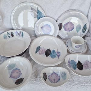 May include: A set of nine white ceramic plates featuring a leaf design in shades of purple, blue, and green. The plates are arranged in a circle on a white surface.