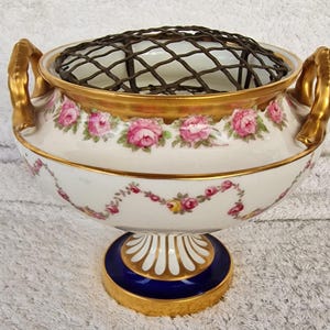 Puede incluir: Un jarrón floral de porcelana blanca con adornos y asas doradas. El jarrón presenta patrones de rosas rosas y una base azul y dorada. Una rejilla de metal oscuro se encuentra dentro.