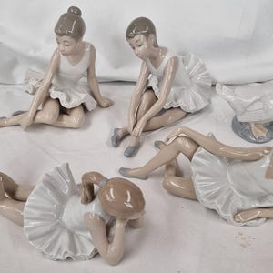 May include: A collection of four porcelain figurines featuring ballet dancers in white tutus and grey ballet slippers, accompanied by a single porcelain goose figurine. Each dancer is depicted in a different seated or reclining pose.