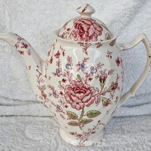 May include: A white ceramic teapot with a lid, featuring a floral pattern in shades of red, pink, green, and blue. The teapot has a curved spout and a handle, suitable for serving tea.