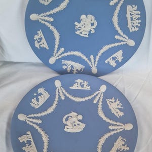 Puede incluir: Dos platos de cerámica azules con diseños en relieve blancos. Cada plato presenta una figura central de querubín y una guirnalda circundante con escenas de relieve clásicas. Los platos son redondos y tienen un acabado mate.