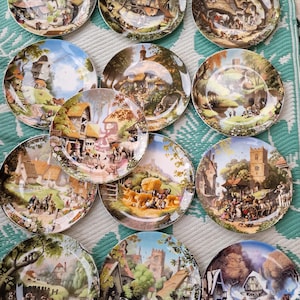 Può includere: Una collezione di 12 piatti in ceramica decorativi che presentano incantevoli scene di campagna. Ogni piatto raffigura un villaggio pittoresco con case, alberi e persone. I piatti sono di vari colori, tra cui verde, marrone, blu e bianco.